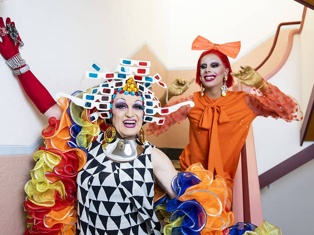 Miss 3D and Etcetera Etcetera Two drag queens pose in a stairwell, Miss 3D wears a headpiece made of retro 3D cardboard glasses and Etcetera Etcetera wears all orange with a big bow on her head.