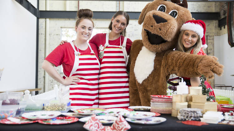 Three people in striped red aprons pose with a giant reindeer.