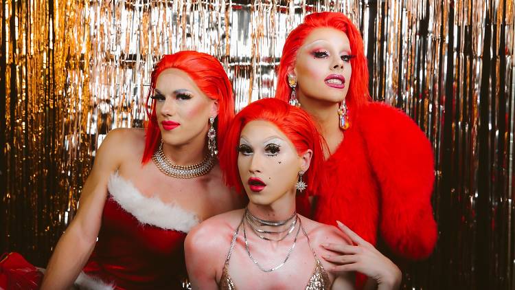 Three drag queens dressed in red festive outfits pose in front of a gold tinsel curtain.