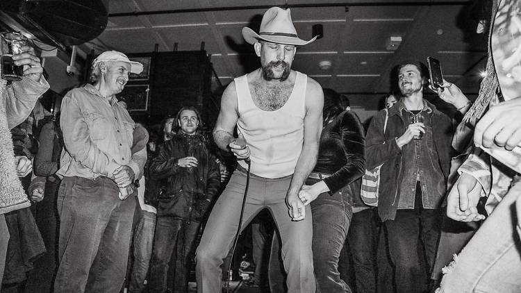 Andy Gollegdge performs amongst a pub crowd, he wears a cowboy hat and a white singlet.