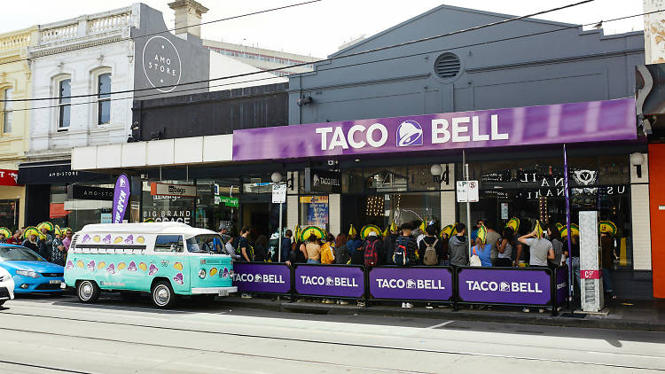 Outside the first taco bell in Victoria. There is a combi van on the street in front of the store and people wearing taco hats lining up outside.