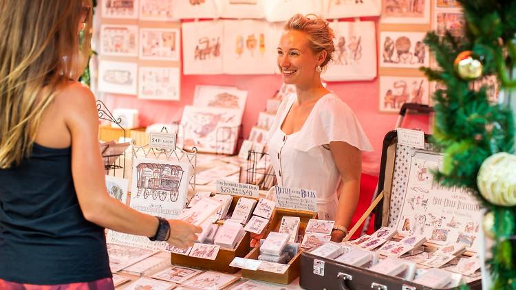 A woman is selling handmade cards and prints in a stall. 