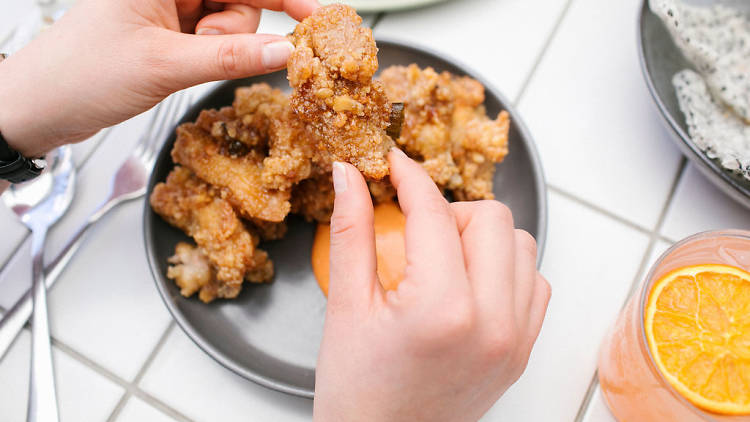 A person's hand hold up a piece of fried chicken from a plate filled with chicken and bright orange sauce.