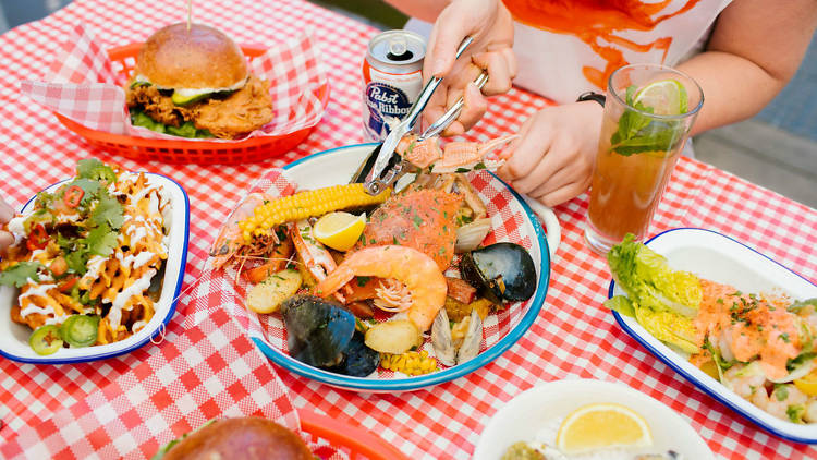 A table covered in dishes including a burger, oysters, loaded fries, wedge salad and a plate of crab boil. Person is cracking one of the crab in the crab boil