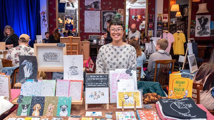 A woman in a head scarf with a blunt fringe poses with a stall of illustrated artworks.