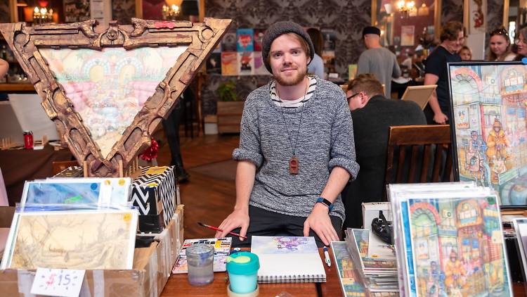 An artist with a beard and beanie poses with a stall of their wares.