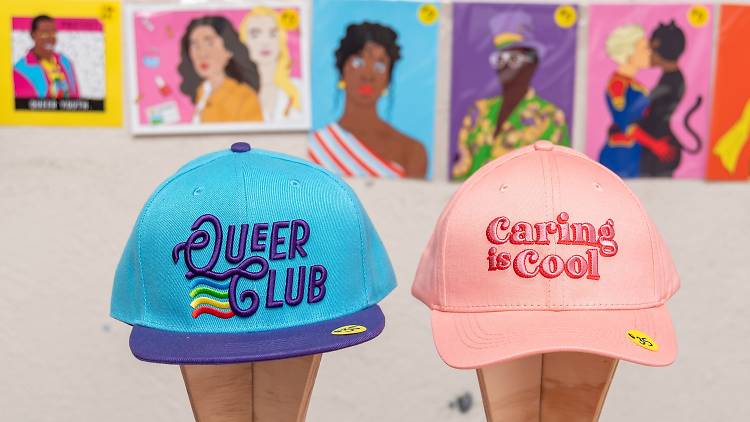 Two baseball caps are on display in front of colourful artwork prints. One is blue and embroidered with the words 'Queer Club', the other is pink and spells 'Caring is Cool'.