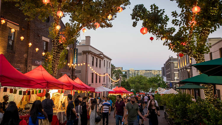 A street lined with market stalls. Overhanging trees are filled with paper lanterns