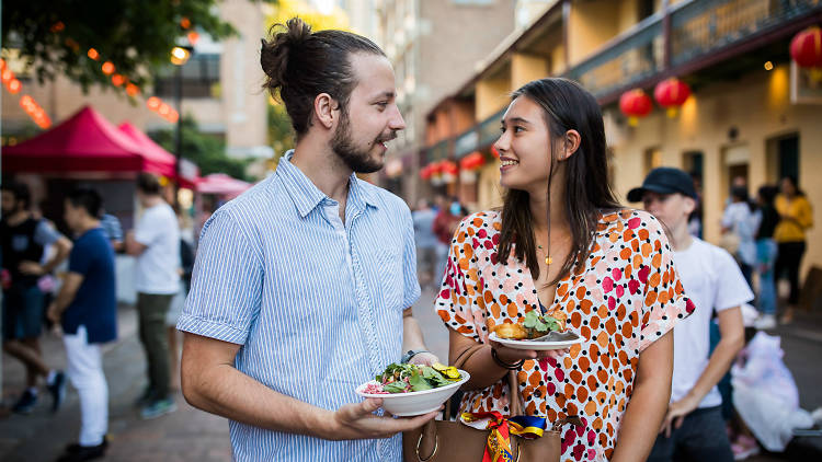 A man and woman standing in the middle of a market, both are holding a plate of food