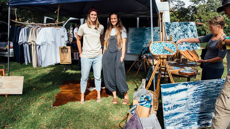 A man and a woman standing outside a market stall filled with clothing. In front of them are ocean-themed artworks