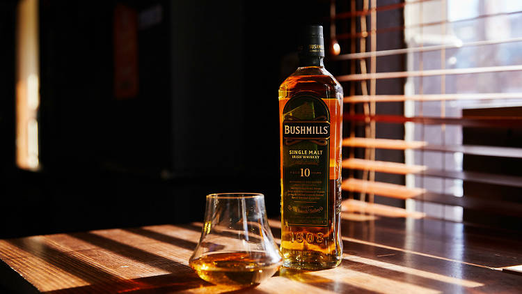 A bottle of Bushmills whiskey and a glass of whiskey next to a window with sunlight streaming in