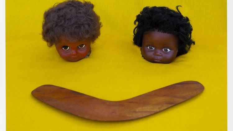 Two doll heads on a yellow backdrop with a wooden boomerang in front of them