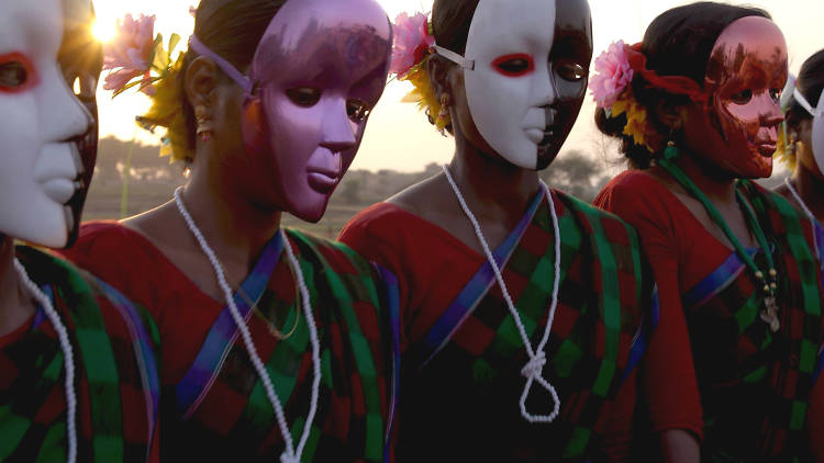 A film still featuring women wearing saris with full-face drama masks over their faces