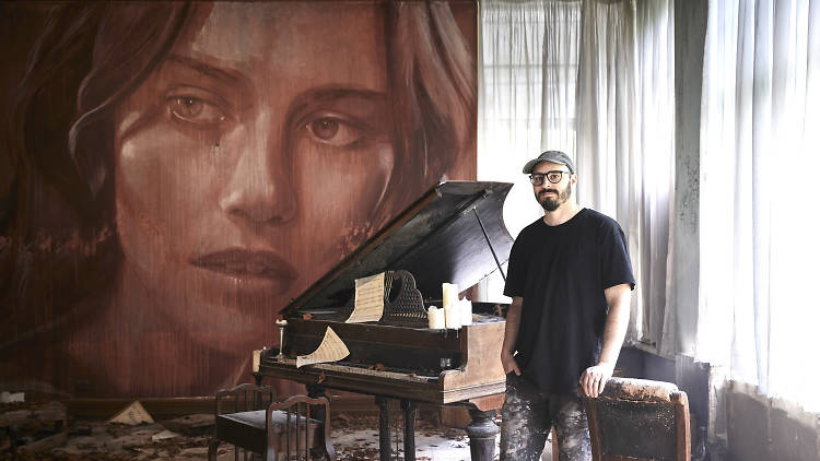 Street artist rone standing next to a baby grand piano and a mural of a woman face 