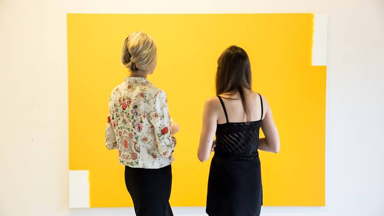 Two women have their backs turned to the camera, they are looking at a large yellow painting. 