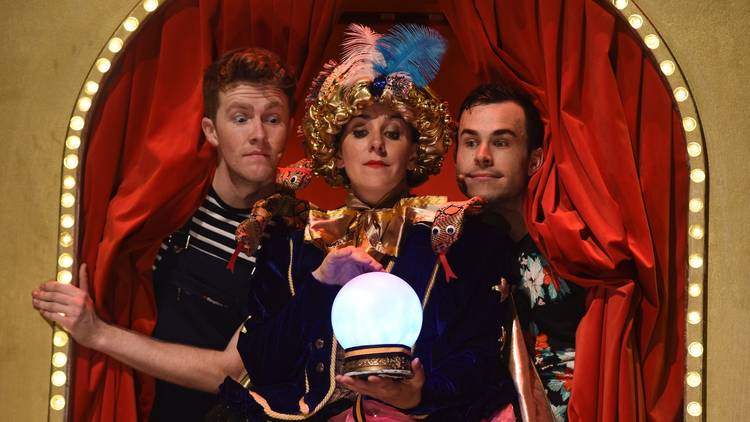 A woman in a fortune teller costume looks over a magic crystal ball as two people peer over her shoulders. 