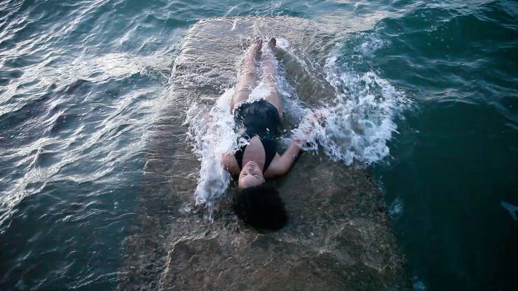 An image of a woman in a black bathing suit lying on a concrete block being covered by ocean waves. 