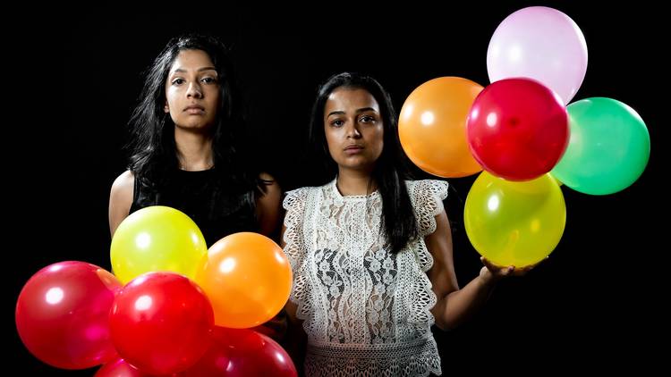 Two women with blank expressions stand holding colourful balloons.