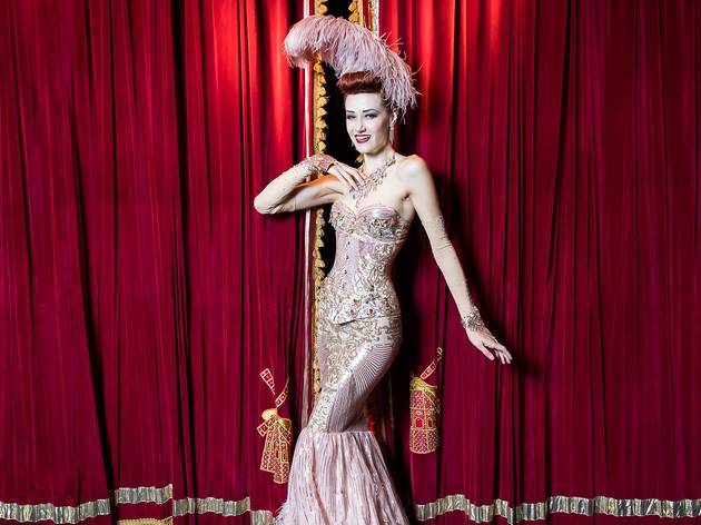 Burlesque in Sydney Porcelain Alice poses in front of a red velvet curtain in a full pink costume with bejewelled corset, skirt and feather head piece.
