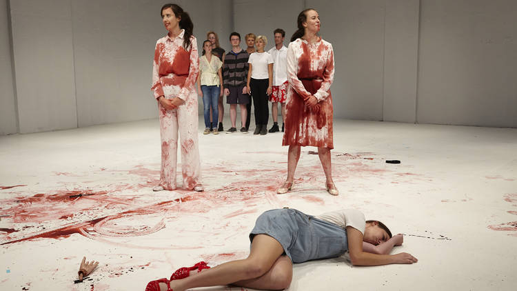 Two women covered in fake blood in a white performance space. A person playing dead lies on the floor in front of them