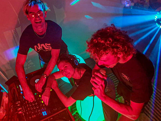 Room 2 Radio Three DJs with a mixing board and disco lights smile up at the camera.