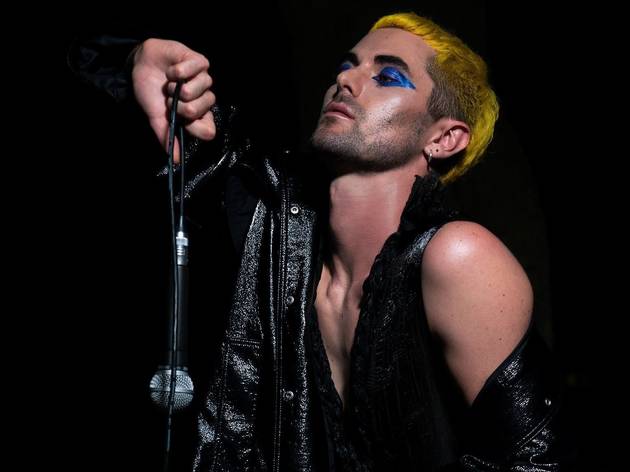 Lavrence for Loud n Queer TV Artist Lavrence performs in all black leather holding a microphone, their cropped hair is bright yellow and they wear blue eyeshadow.