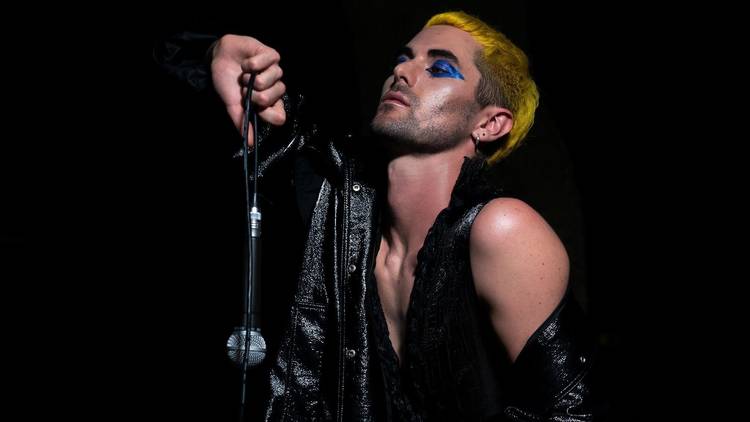 Artist Lavrence performs in all black leather holding a microphone, their cropped hair is bright yellow and they wear blue eyeshadow.