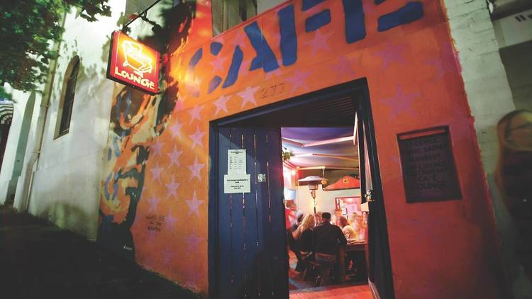 The doorway of Cafe Lounge, a painted blue wooden door opens with a neon sign overhead.