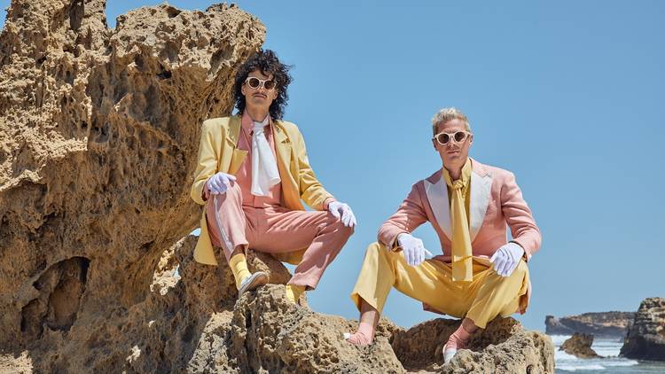 Two men in pink and yellow pastel suits pose on a rocky beach.