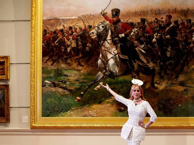 Verushka Darling AGNSW tour A Drag queen poses in front of a large painting depicting an army on horseback.