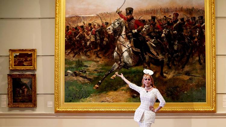 A Drag queen poses in front of a large painting depicting an army on horseback.