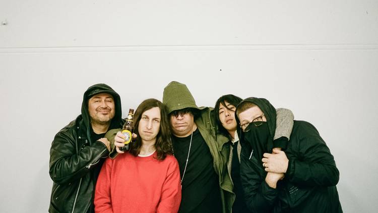 Band poses, huddled together in front of a blank wall. one holds a beer.