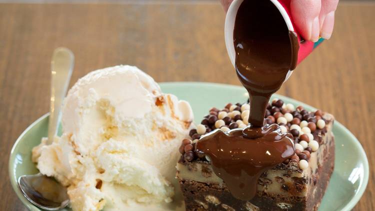 A brownie on a plate next to a scoop of ice cream. A shot of melted chocolate is being poured over the brownie