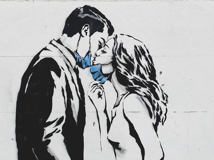 A graffiti painting of a man and woman kissing, they have blue medical masks around their chins.