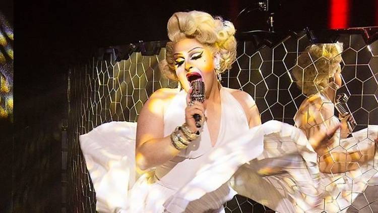 Drag queen dressed as Marilyn Monroe in white dress and blonde wig sings into microphone.