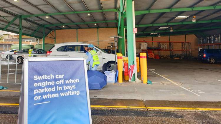 staff in medical gear speak to people in a parked car, a sign reads: 'Switch car engine off when parked in bays or when waiting'.