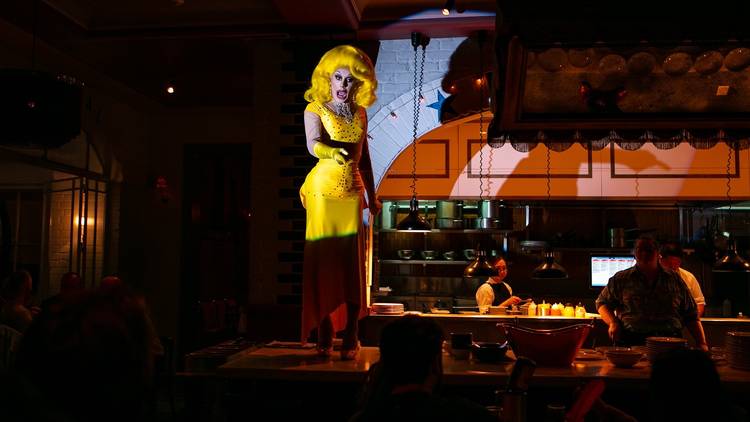 Drag queen performs in yellow dress and gloves, she stands on a restaurant table