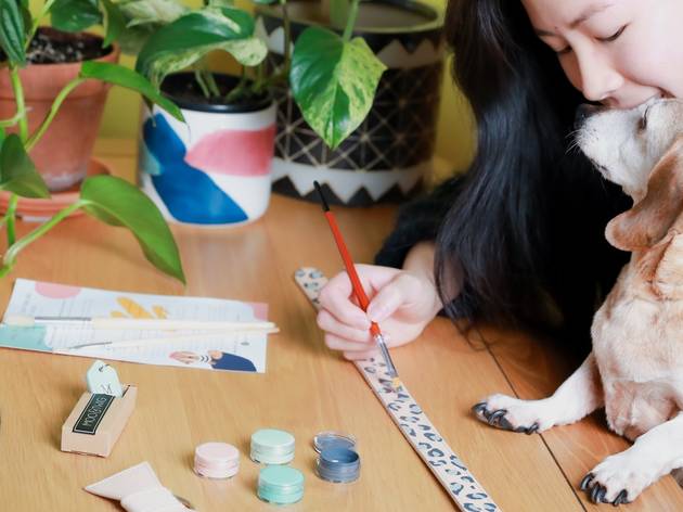 Moo + Twig DIY dog collar kit A woman and dog sit at a table, she is painting a collar, there are craft supplies on the table.
