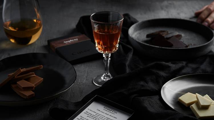 A dark table featuring plates of chocolate and dainty glasses filled with alcohol