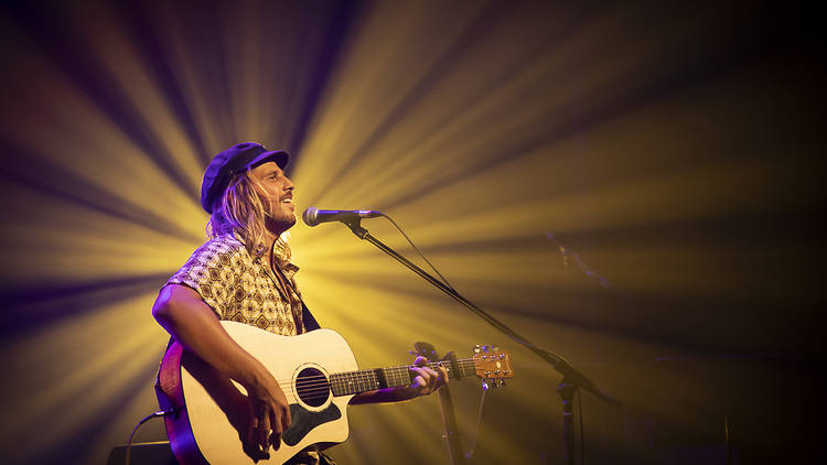 Singer Kyle Lionheart sitting on stage with a microphone and a acoustic guitar. A starburst of golden light is behind him