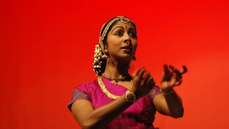 A dancer in a pink sari and gold jewellery performs against a bright red backdrop 