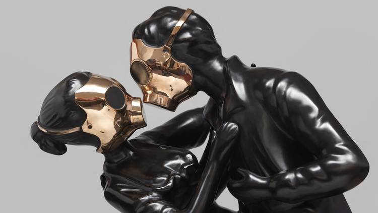 Two sculptural figures in matte black wear bronze gas masks and embrace