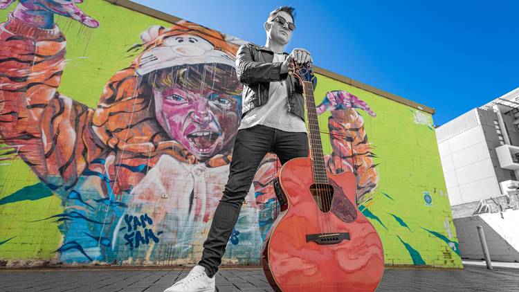 Former INXS lead singer Ciaran Gribbin poses with a guitar in front of bright graffiti