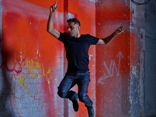 New Breed 2020 A man in black t-shirt and jeans jumps with one finger in the air in front of a red spray-painted warehouse brick wall