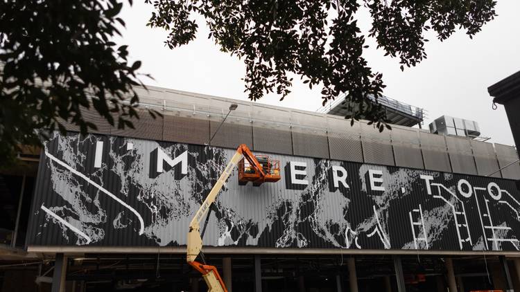 a giant mural going up with artist Georgia Hill in a cherry picker. This section reads I'm Here Too