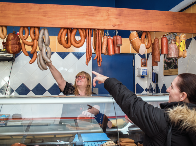 Two ladies in a deli. One pointing at cured sausages, other being the deli assistant picking it out for her.