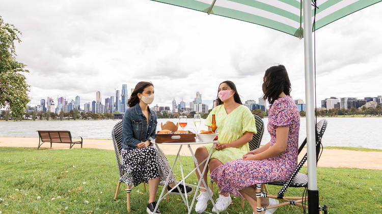 Three ladies sitting on chairs by a table with wine, beers and food under an umbrella. View behind is the city's skyline and lake.