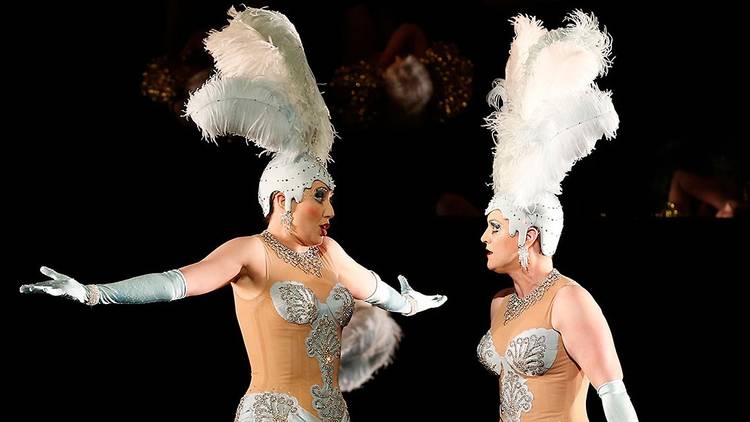 Two performers wear bejwelled light blue costumes with feathered head pieces.