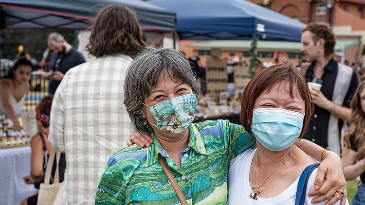 Two people wearing face masks smile with their arms around each other, they are at a market.