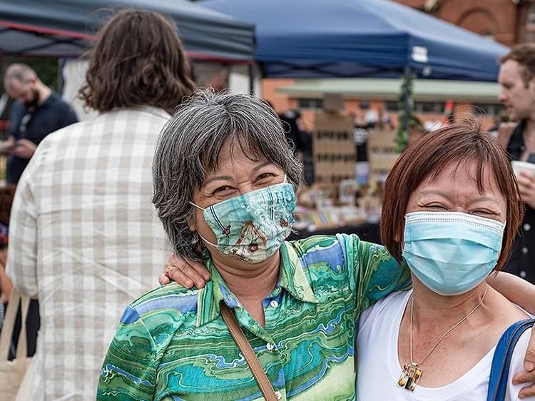 Two people wearing face masks smile with their arms around each other, they are at a market.